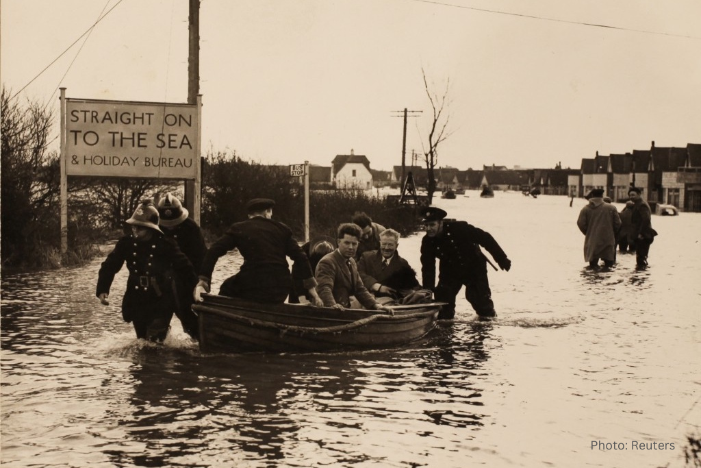 Photo of people in a boat and wading through water along a flooded road with a sign in the background saying To the sea, credit Reuters