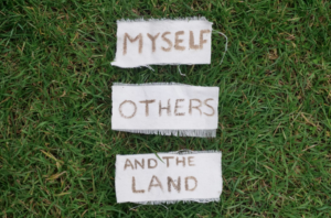 Text saying Myself Others and the Land painted onto three pieces cloth laid out on green grass